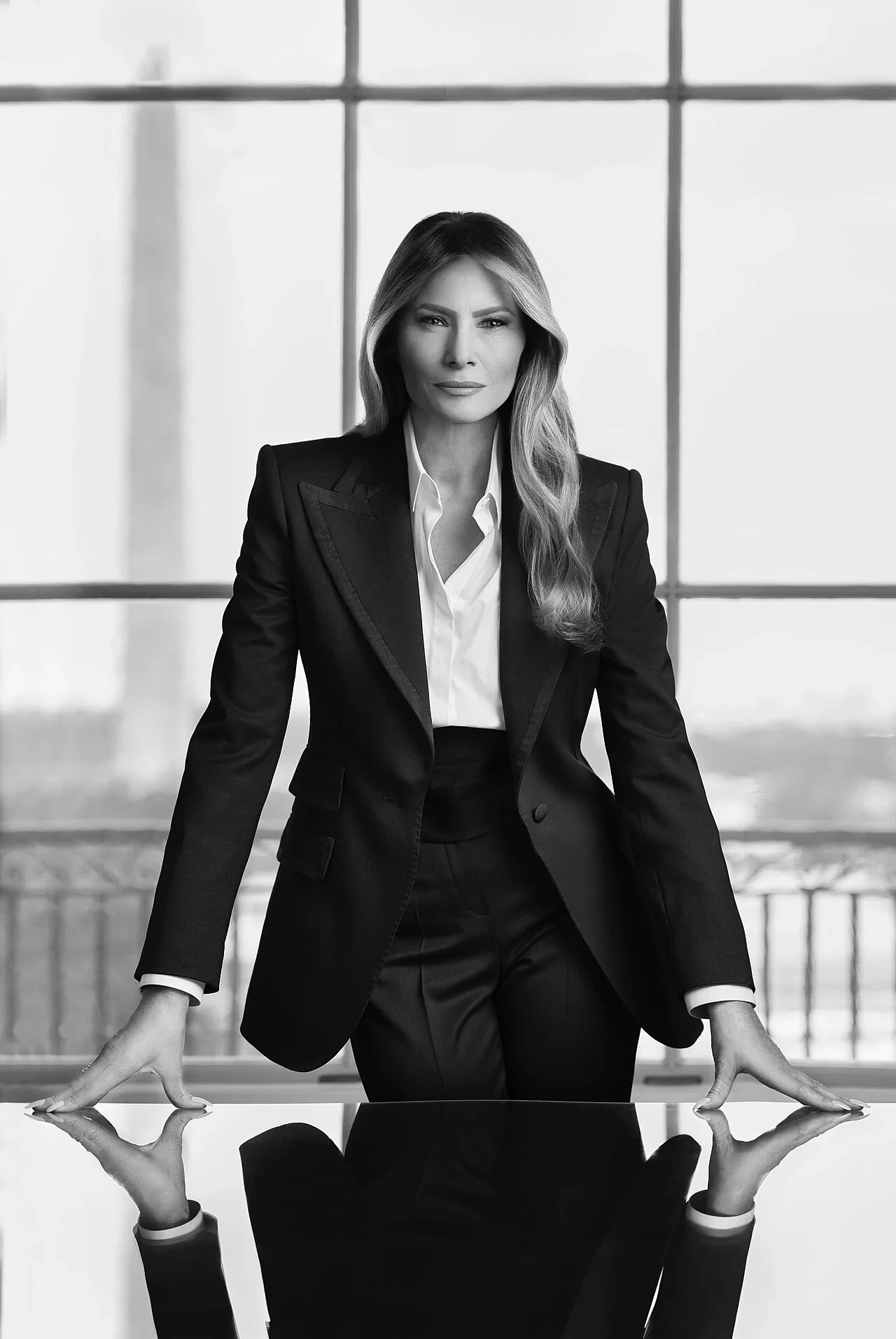 File photo, taken in January 27, 2025: Melania Trump stands in a dark suit and white shirt in front of a window and a reflective table.