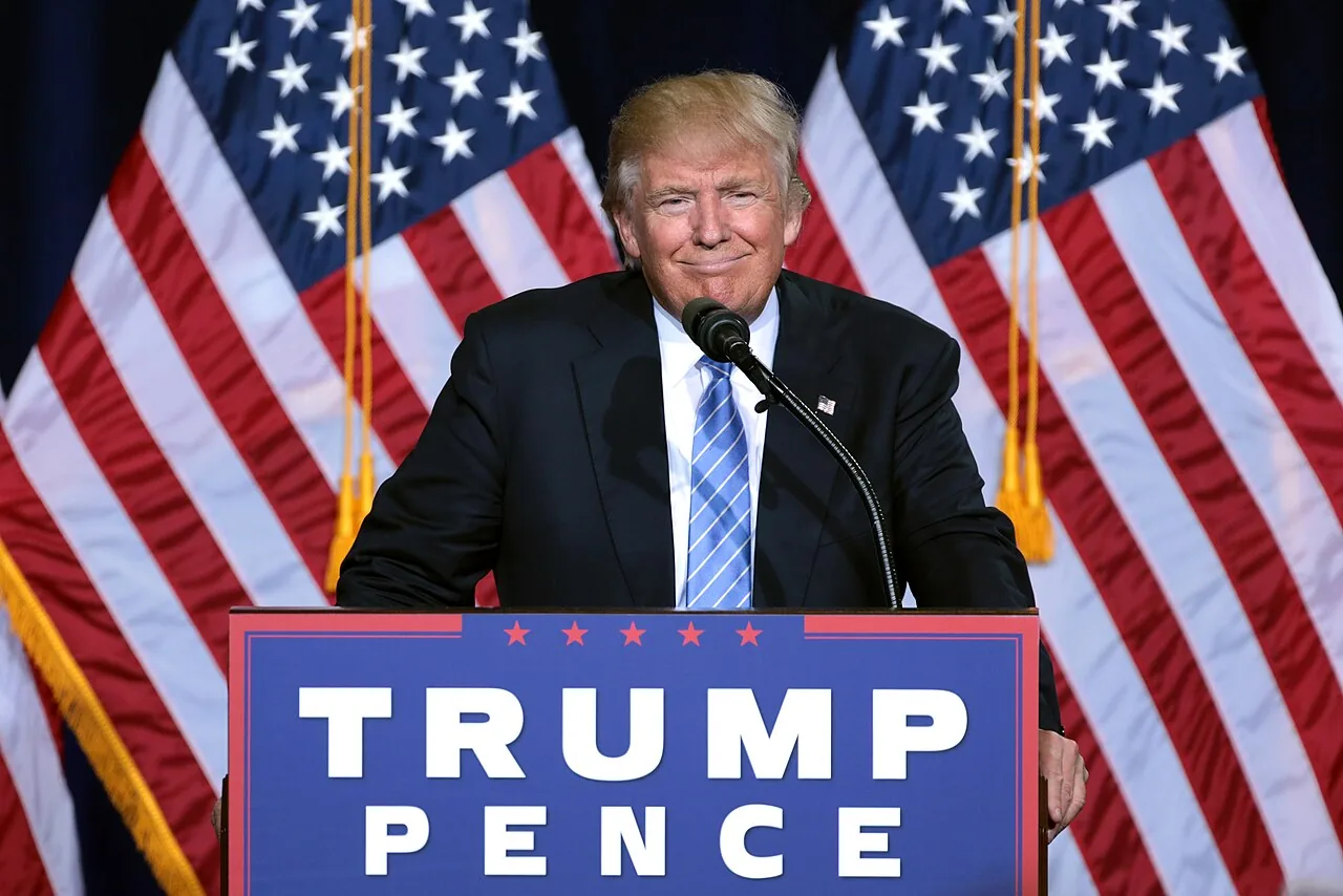 File photo from 31 August 2016: Donald Trump speaks into a microphone behind a lectern bearing the words “Trump Pence,” with U.S. flags in the background.